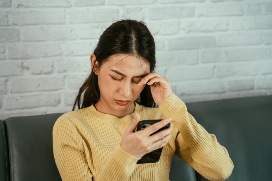 A person looking down at a phone and rubbing their eye.
